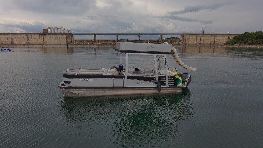 Gray Avalon pontoon boat with upper deck slide anchored near dam wall on the lake.