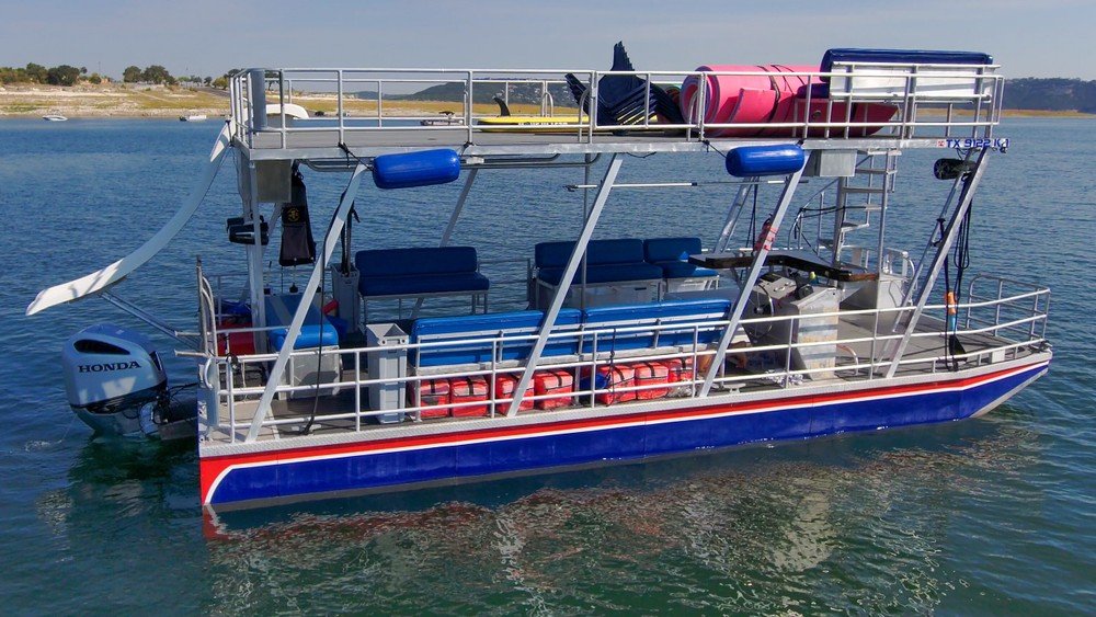 Blue double-decker pontoon boat with slide and floating mats, anchored on Lake Travis.
